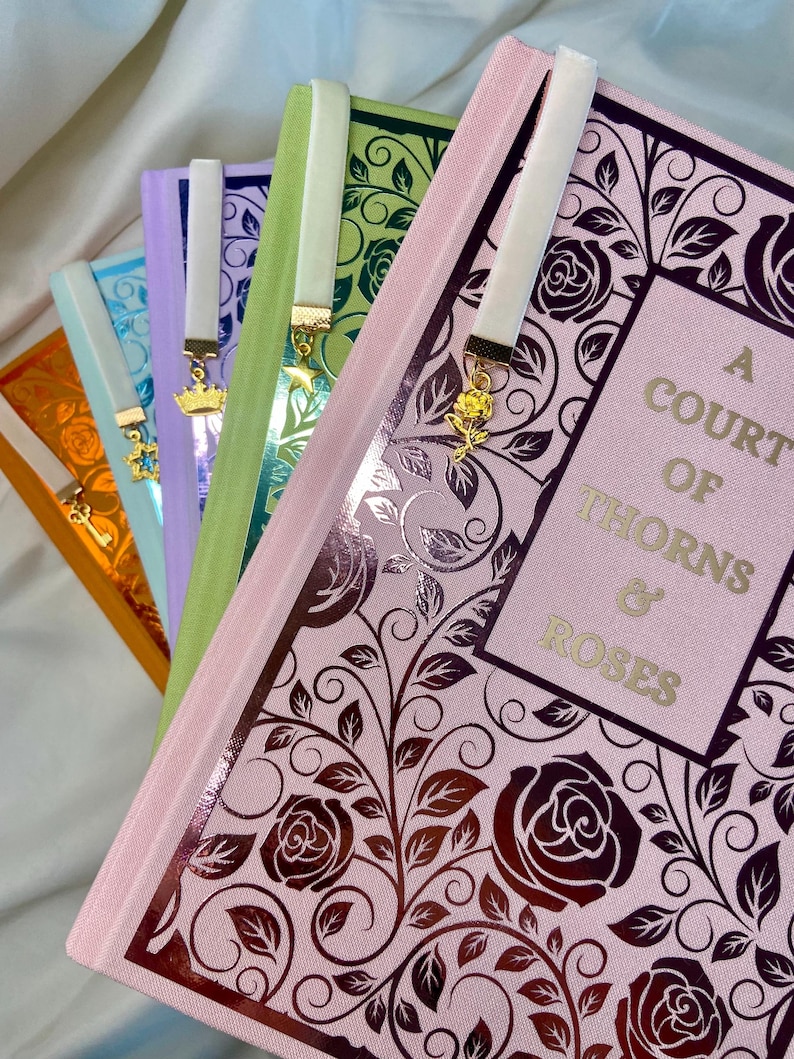 Pode incluir: Um conjunto de cinco livros com marcadores de fita de cetim combinando. Os livros s&atilde;o de cores diferentes e t&ecirc;m um design floral na capa. O livro de cima &eacute; rosa e tem o t&iacute;tulo "A Court of Thorns & Roses" na capa.