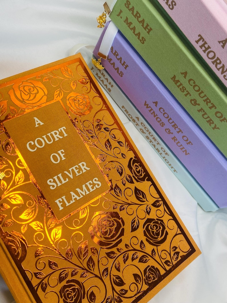 Pode incluir: A Court of Silver Flames de Sarah J. Maas. A capa do livro apresenta um design floral dourado e laranja com um t&iacute;tulo em texto branco.