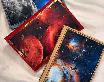 以下が含まれることがあります： 宇宙をテーマにした3冊のハードカバー本。「Artemis」、「The Martian」、「Project Hail Mary」というタイトルで、Andy Weir著。表紙には、惑星、星雲、宇宙飛行士の画像が星空を背景に描かれています。本は金色の縁取りがされています。