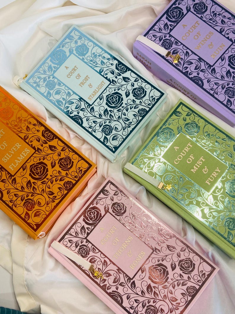 Pode incluir: Cinco livros de capa dura com desenhos florais e letras douradas. Os t&iacute;tulos s&atilde;o "A Court of Thorns and Roses", "A Court of Mist and Fury", "A Court of Wings and Ruin", "A Court of Frost and Starlight", e "A Court of Silver Flames".