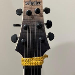 Op de afbeelding: Close-up van een gitaarkop met de merknaam "Schecter" en "Diamond Series" erop gedrukt. De kop heeft zwarte stemknoppen en een gradiëntafwerking. Een gele gehaakte gitaarcapo zit op de hals.