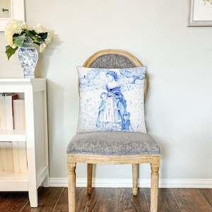 May include: A gray upholstered chair with wooden legs and a white and blue pillow featuring a watercolor illustration of a woman and two children in a garden setting.