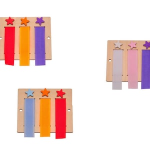 Op de afbeelding: Drie houten planken met stervormige uitsnijdingen en gekleurde stofstroken eraan bevestigd. De planken zijn in een rij gerangschikt, waarbij de bovenste plank rode, oranje en paarse stroken heeft, de middelste plank witte, roze en paarse stroken heeft en de onderste plank blauwe, oranje en rode stroken heeft.