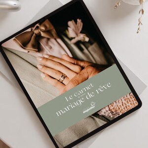 Peut inclure: Une tablette numérique affichant un graphique vert et blanc avec le texte "Le carnet de notre mariage de rêve" et le logo "Les Six Magnolias". L'image sur la tablette montre un gros plan des mains d'un couple avec une alliance en or.