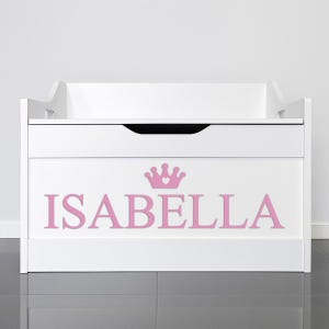 May include: A white wooden toy chest with a lift-up lid and a lower drawer. The name "ISABELLA" is displayed in pink letters, topped with a pink crown graphic. The chest is designed for storage.