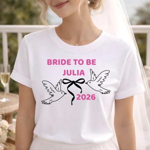 Könnte beinhalten: Weißes T-Shirt mit dem Text "BRIDE TO BE JULIA 2026" in Pink. Unter dem Text sind zwei weiße Tauben mit schwarzen Umrissen abgebildet, die durch eine schwarze Schleife verbunden sind.