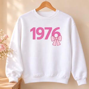 Könnte beinhalten: Ein weißes langärmeliges Sweatshirt mit Rundhalsausschnitt und dem rosafarbenen Text 1976 auf der Vorderseite. Die Zahl Sechs ist mit einer rosafarbenen Schleifengrafik verziert. Das Kleidungsstück hängt auf einem Holzkleiderbügel.