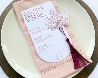May include: A white arched menu card with the text EAT, DRINK AND BE MARRIED, STARTER, ENTREE, and DESSERT, featuring a maroon orchid-shaped name tag for Henry and a matching maroon tassel, resting on a pale pink napkin atop a white plate.