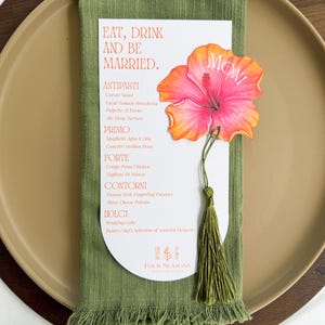 Op de afbeelding: Een witte, boogvormige menukaart met de tekst Eat, Drink and be Married, voorzien van een oranje en roze hibiscusbloem-uitsnede met een groene kwast. De menukaart ligt op een olijfgroene servet op een beige bord.