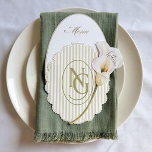 May include: An oval menu card with the word Menu and a scalloped beige and white striped insert featuring the letters NG in a central oval. A white calla lily cutout with the name Katherine is attached to the right side, resting on a sage green napkin.