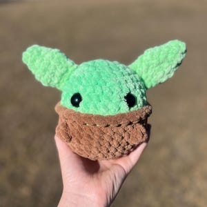 Puede incluir: Un juguete de peluche hecho a mano con una cabeza verde, orejas grandes y ojos negros, colocado en un portabeb&eacute;s tejido marr&oacute;n. El juguete se sostiene en una mano, destacando la textura suave y la artesan&iacute;a detallada.