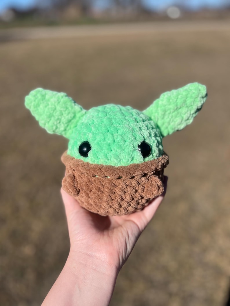 Puede incluir: Un juguete de peluche hecho a mano con una cabeza verde, orejas grandes y ojos negros, colocado en un portabeb&eacute;s tejido marr&oacute;n. El juguete se sostiene en una mano, destacando la textura suave y la artesan&iacute;a detallada.