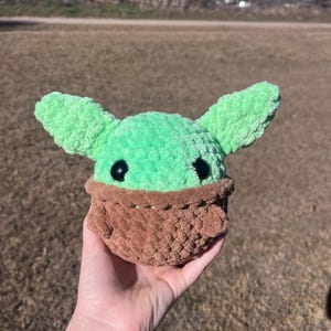 Puede incluir: Un juguete de peluche hecho a mano de un personaje con orejas grandes, cabeza verde y ropa marr&oacute;n. El juguete tiene ojos negros y se sostiene en una mano contra un fondo borroso al aire libre. El peluche est&aacute; hecho de hilo suave y texturizado.