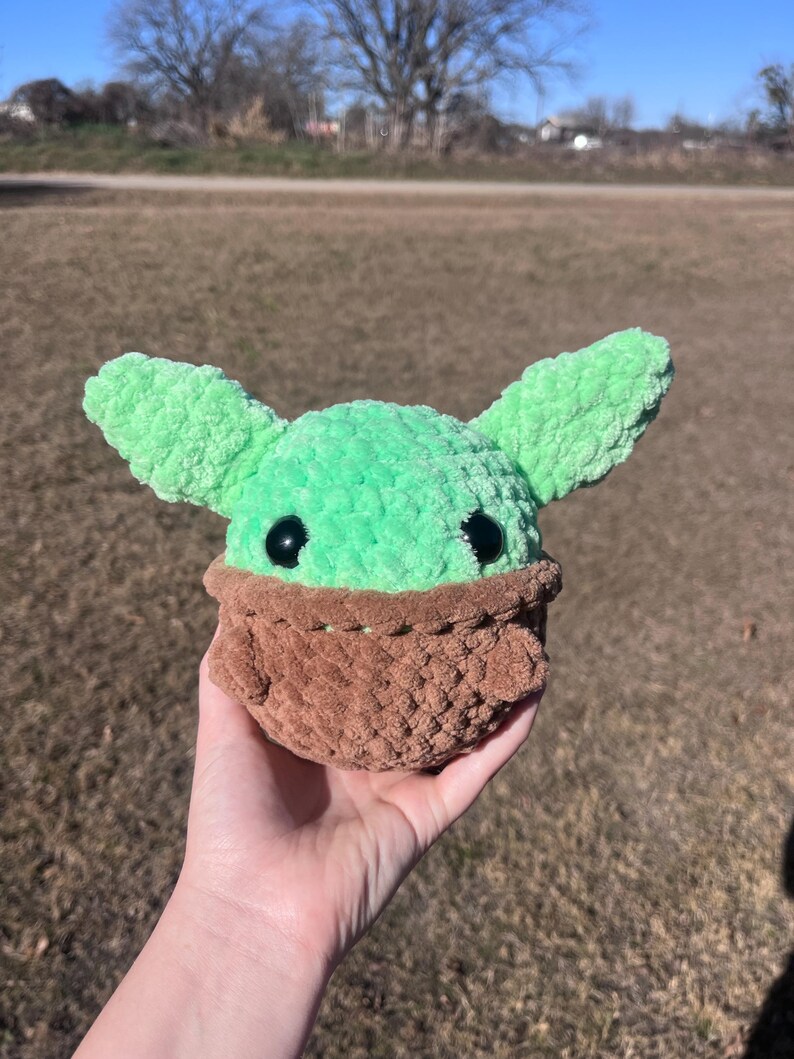 Puede incluir: Un juguete de peluche hecho a mano de un personaje con orejas grandes, cabeza verde y ropa marr&oacute;n. El juguete tiene ojos negros y se sostiene en una mano contra un fondo borroso al aire libre. El peluche est&aacute; hecho de hilo suave y texturizado.