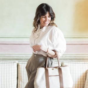 May include: A woman holding a large tote bag. The bag is white with brown leather trim and handles. She is wearing a white blouse and gray pants. The background is a light green and pink wall.