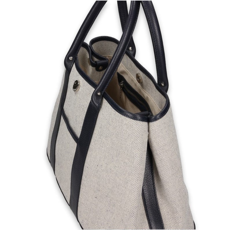 Pu&ograve; includere: Una borsa tote beige con finiture e manici in pelle blu navy. La borsa ha un design strutturato con una tasca interna con cerniera e una tasca laterale. I manici sono fissati con hardware color argento.
