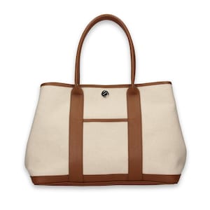 Pu&ograve; includere: Borsa tote color crema con finiture e manici in pelle marrone. La borsa ha una tasca frontale e una chiusura in metallo argentato. I manici sono arcuati e la borsa ha una forma rettangolare.