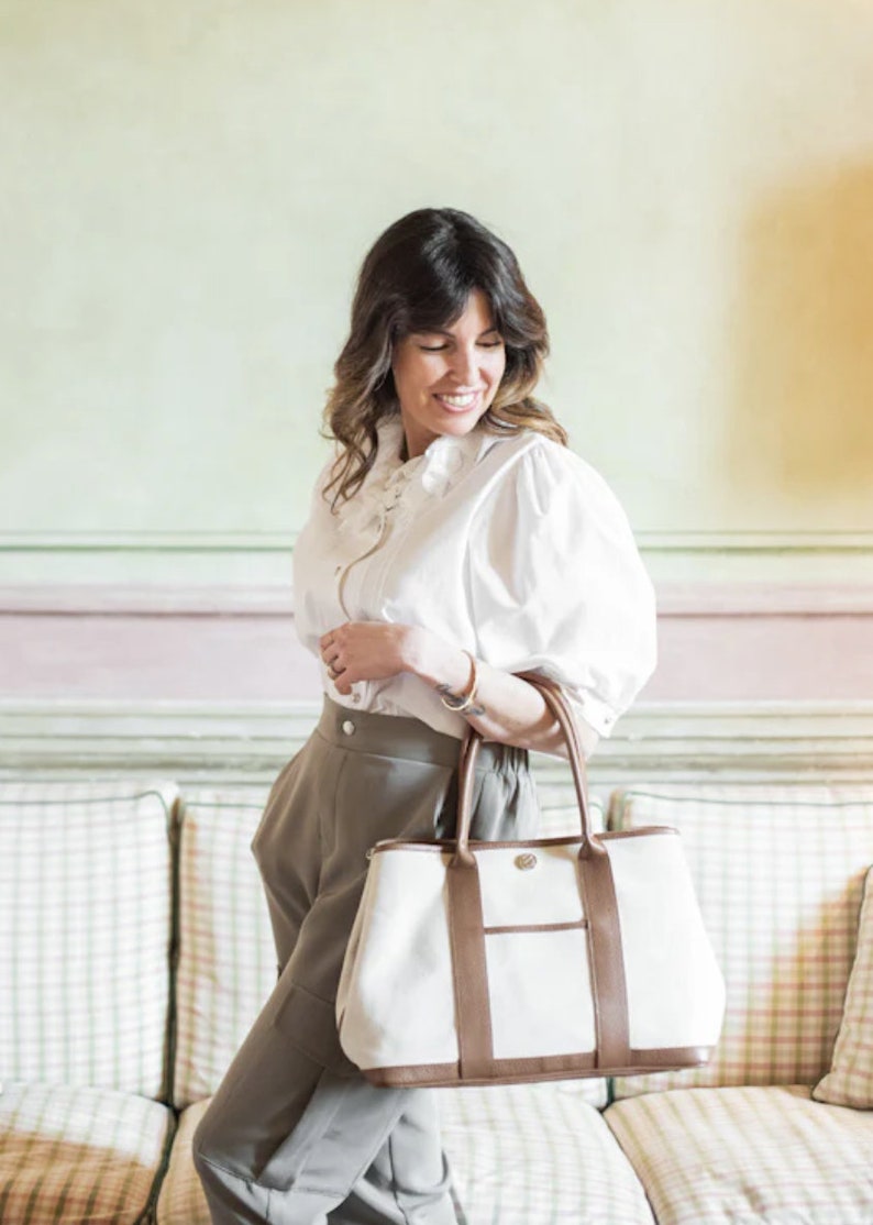 May include: A woman holding a large tote bag. The bag is white with brown leather trim and handles. She is wearing a white blouse and gray pants. The background is a light green and pink wall.