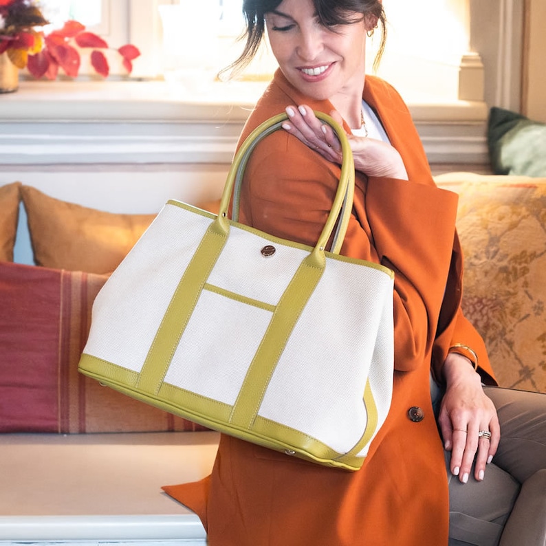 Pu&ograve; includere: Una grande borsa tote con corpo in tela color crema e finiture in pelle verde oliva. La borsa ha una forma rettangolare, una tasca frontale e due manici superiori. Una chiusura in metallo color argento &egrave; visibile sulla parte anteriore.