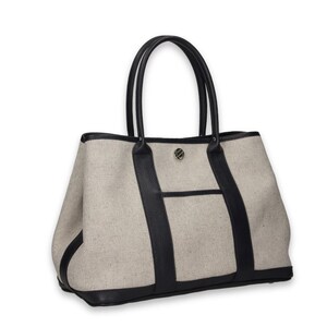 Pu&ograve; includere: Borsa tote beige con dettagli in pelle nera. La borsa presenta manici neri, base nera e finiture nere. Una piccola tasca &egrave; visibile sul davanti e un'emblema argentato &egrave; centrato sul davanti.