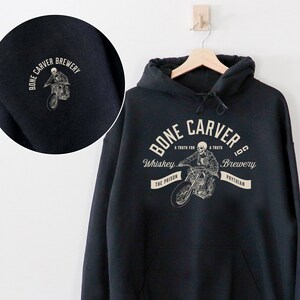 Könnte beinhalten: Ein schwarzer Kapuzenpullover mit einem Aufdruck auf der Vorderseite mit dem Text "BONE CARVER BREWERY" und einem Skelett auf einem Motorrad. Der linke Ärmel hat ein kleineres Brauerei-Logo. Der Pullover hängt an einem Holzkleiderbügel.