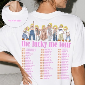 May include: A white t-shirt featuring a graphic of eight cartoon figures in various outfits above the pink text "the lucky me tour" and a list of tour dates. The front neckline area displays the pink text "sing to me hilary" in a small circle.