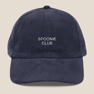 Op de afbeelding: Een donkerblauwe corduroy baseballpet met witte geborduurde tekst die "SPOONIE CLUB" luidt.