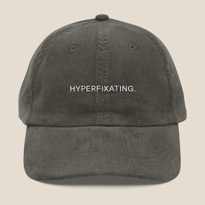May include: An olive green corduroy baseball cap with a curved peak. The cap displays the white text "HYPERFIXATING." across the front. The hat has a structured crown and a small button on top.