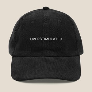Puede incluir: Una gorra de béisbol de pana negra con texto bordado en blanco que dice "OVERStimulated".