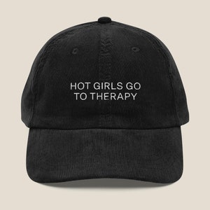 Op de afbeelding: Zwarte corduroy baseballpet met witte geborduurde tekst die "HOT GIRLS GO TO THERAPY" zegt.