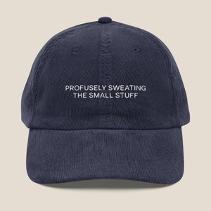 May include: A navy blue corduroy baseball cap with white embroidery that reads "PROFUSELY SWEATING THE SMALL STUFF".