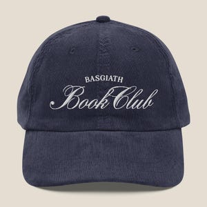 以下が含まれることがあります： 紺色のコーデュロイ野球帽。「BASGIATH Book Club」の文字が白い筆記体で刺繍されています。カーブしたつばと、後ろに調節可能なストラップが付いています。