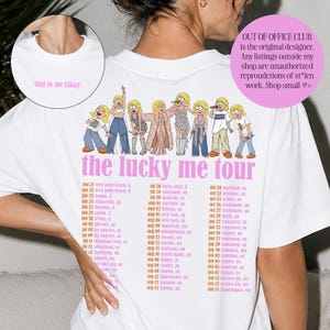 Op de afbeelding: Een wit t-shirt met een roze afbeelding op de achterkant met acht stripfiguren, de tekst "the lucky me tour" en een lijst met tourdata. De voorkant bij de hals toont de kleine roze tekst "sing to me hilary" in een cirkelvormig inzetstuk.