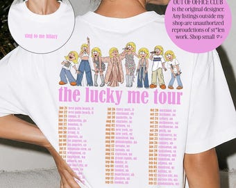 Pode incluir: Uma t-shirt branca com um gráfico cor-de-rosa nas costas com oito figuras de desenhos animados, o texto "the lucky me tour" e uma lista de datas da digressão. A zona do decote frontal apresenta o pequeno texto rosa "sing to me hilary" num círculo.