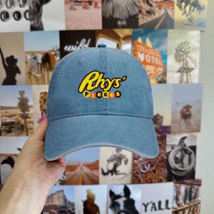 Peut inclure: Casquette de baseball en denim bleu clair avec le texte "Rhys' Pieces" en jaune et orange. La casquette a une visière incurvée et une conception classique à six panneaux. Un accessoire décontracté.