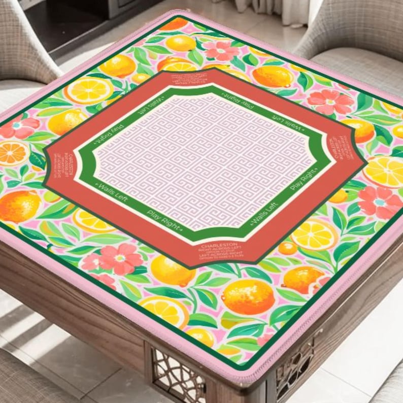 May include: A square table cover with a vibrant fruit and floral design. The cover features lemons, orange slices, and pink flowers on a light pink background. A central geometric design is framed by a red and green border with text.