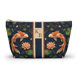 May include: A zippered pouch featuring a koi fish design. The pouch has a dark blue background with koi fish, lotus flowers, and foliage. The center stripe has the letters "K" and "B".