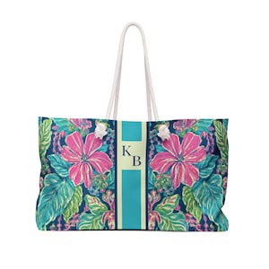 May include: Large tote bag with a floral pattern featuring pink flowers and teal leaves on a navy blue background. The bag has a teal stripe down the center with the initials "K B" and white rope handles.