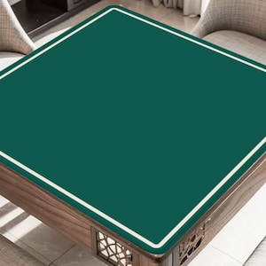 Könnte beinhalten: Ein quadratischer Holztisch mit einer dunkelgrünen Oberfläche und einem weißen Rand. Der Tisch hat einen dekorativen Holzsockel mit aufwendigen Ausschnitten. Der Tisch ist von zwei Stühlen umgeben.