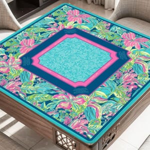 以下が含まれることがあります： 鮮やかな花柄の正方形のテーブルカバー。カバーは、ティールブルーの縁、ネイビーブルーの中央、ピンクの縁取りのある中央のターコイズダイヤモンド型が特徴です。花柄にはピンクと緑の花が含まれています。