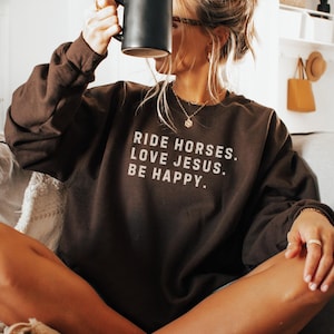 May include: Chocolate-colored sweatshirt with the text "RIDE HORSES. LOVE JESUS. BE HAPPY." in white. The sweatshirt is worn with a black mug. The color of the sweatshirt is labeled "chocolate".