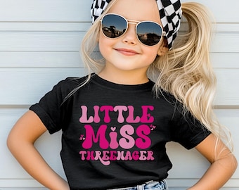 May include: A black short-sleeve t-shirt featuring the pink and light pink text Little Miss Threenager with small heart accents. The shirt is paired with blue denim shorts, a black and white checkered hair bow, and dark-tinted sunglasses.