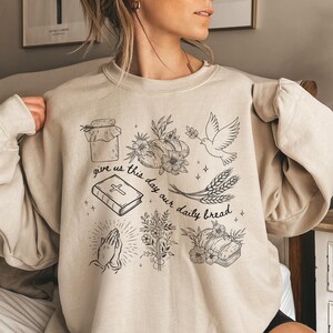 May include: Sand-colored sweatshirt with a black graphic design featuring a jar, flowers, a dove, wheat, a book, praying hands, and bread. The text "give us this day our daily bread" is also included. The sweatshirt is a neutral color.
