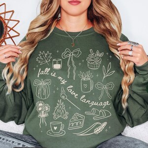 May include: A military green crewneck sweatshirt featuring a cream-colored line art graphic with the text "fall is my love language" surrounded by illustrations of a maple leaf, candle, cupcake, pumpkin, mug, bows, campfire, s'more, flowers, slippers, and pie.