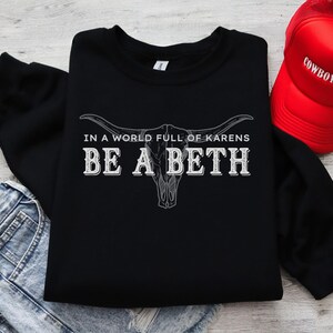 May include: Black sweatshirt with the text "IN A WORLD FULL OF KARENS BE A BETH" in white, along with a graphic of a bull skull. A red trucker hat with "COWBOY HAT" is visible in the upper right corner. The sweatshirt is folded.