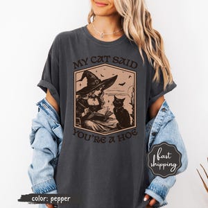 May include: Pepper-colored t-shirt featuring a vintage-style illustration of a witch reading a book with a black cat. The text on the shirt reads "MY CAT SAID YOU'RE A HOE". The design includes bats and a witch hat.
