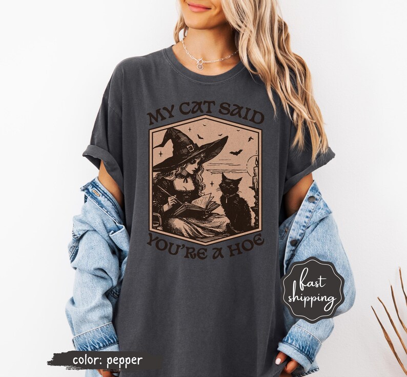 May include: Pepper-colored t-shirt featuring a vintage-style illustration of a witch reading a book with a black cat. The text on the shirt reads "MY CAT SAID YOU'RE A HOE". The design includes bats and a witch hat.