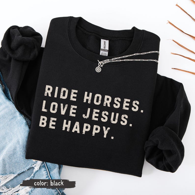 May include: Black sweatshirt with the text "RIDE HORSES. LOVE JESUS. BE HAPPY." in a distressed font. A silver necklace with a circular pendant is draped across the sweatshirt. The sweatshirt is folded on top of blue jeans.