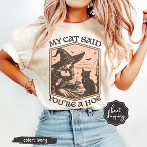 May include: Ivory-colored t-shirt featuring a vintage-style illustration of a witch reading a book with a black cat. The text reads "MY CAT SAID YOU'RE A HOE". The shirt has short sleeves and is worn with blue jeans.