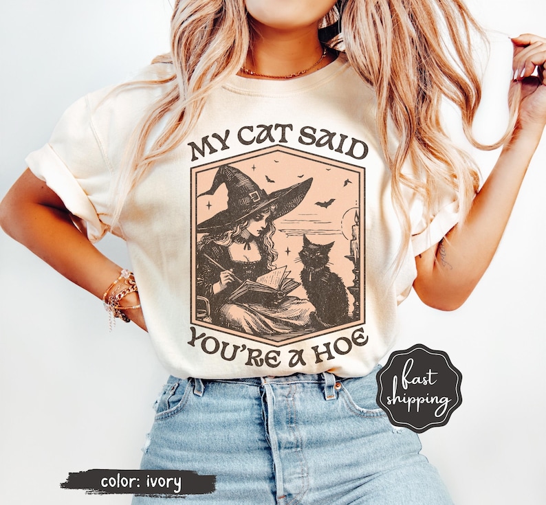 May include: Ivory-colored t-shirt featuring a vintage-style illustration of a witch reading a book with a black cat. The text reads "MY CAT SAID YOU'RE A HOE". The shirt has short sleeves and is worn with blue jeans.